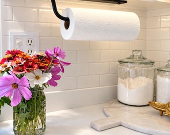 May include: A matte black metal paper towel holder mounted underneath a white kitchen cabinet, holding a roll of white paper towels. The holder features a horizontal bar with a curved arm that supports the roll, providing a functional storage solution.