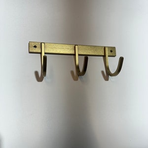 May include: Gold metal wall mounted hook rack with three hooks. The hooks are curved and have a brushed finish.