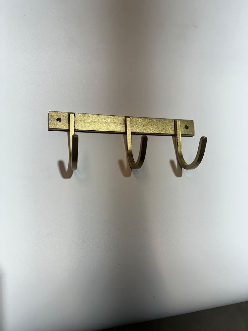 May include: Gold metal wall mounted hook rack with three hooks. The hooks are curved and have a brushed finish.