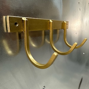 May include: Gold metal wall mounted hook rack with three hooks. The hooks are in the shape of a U.