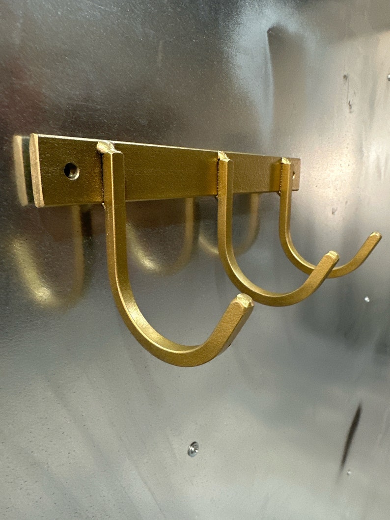 May include: Gold metal wall mounted hook rack with three hooks. The hooks are in the shape of a U.