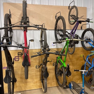 May include: Five bicycles are mounted on a wall using a wall-mounted bike rack system. The bikes are a mix of colors, including black, red, green, purple, and blue. The bikes are all mountain bikes with knobby tires.