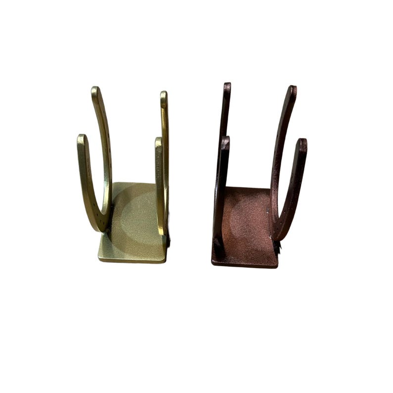 May include: Two U-shaped metal holders, one in a brushed gold finish and one in a dark bronze finish. Each holder features a flat rectangular base and two vertical curved arms that resemble the shape of a horseshoe, standing upright.