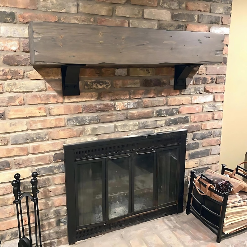 May include: A rustic wooden mantel shelf with black metal brackets mounted above a black fireplace with glass doors. The fireplace is set against a brick wall.