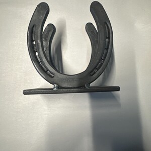 May include: A dark gray metal holder constructed from two U-shaped horseshoes welded together in an upright position. The horseshoes are attached to a flat rectangular metal base plate, featuring visible nail holes and stamped text on the metal surface.