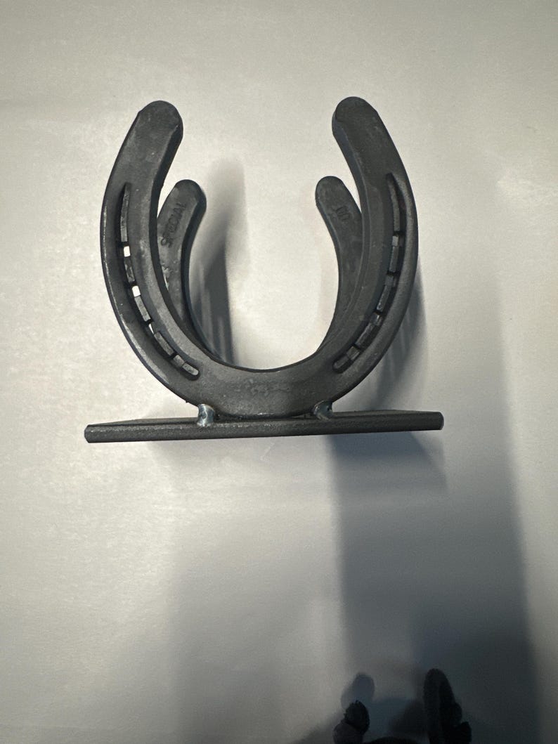 May include: A dark gray metal holder constructed from two U-shaped horseshoes welded together in an upright position. The horseshoes are attached to a flat rectangular metal base plate, featuring visible nail holes and stamped text on the metal surface.