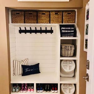 K&ouml;nnte beinhalten: Eine wei&szlig;e Einbau-Garderobe mit f&uuml;nf geflochtenen K&ouml;rben im oberen Fach, einer schwarzen Garderobenleiste mit acht Haken und einer Sitzbank. Ein gestreiftes Kissen und ein marineblaues Kissen mit dem Text This is home liegen auf der Sitzbank.