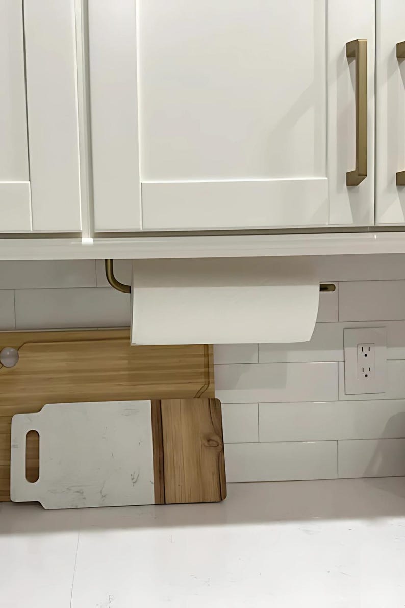 Puede incluir: Un porta rollo de papel de cocina blanco con un brazo de metal dorado montado debajo de un armario de cocina blanco. El porta rollo tiene un rollo de papel de cocina blanco. Hay una tabla de cortar de madera en la encimera debajo del porta rollo.