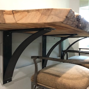 May include: A live edge wood countertop with two black metal brackets attached to a white wall. The brackets are curved and have a decorative design. The countertop is supported by the brackets and is ready to be used as a table or bar.