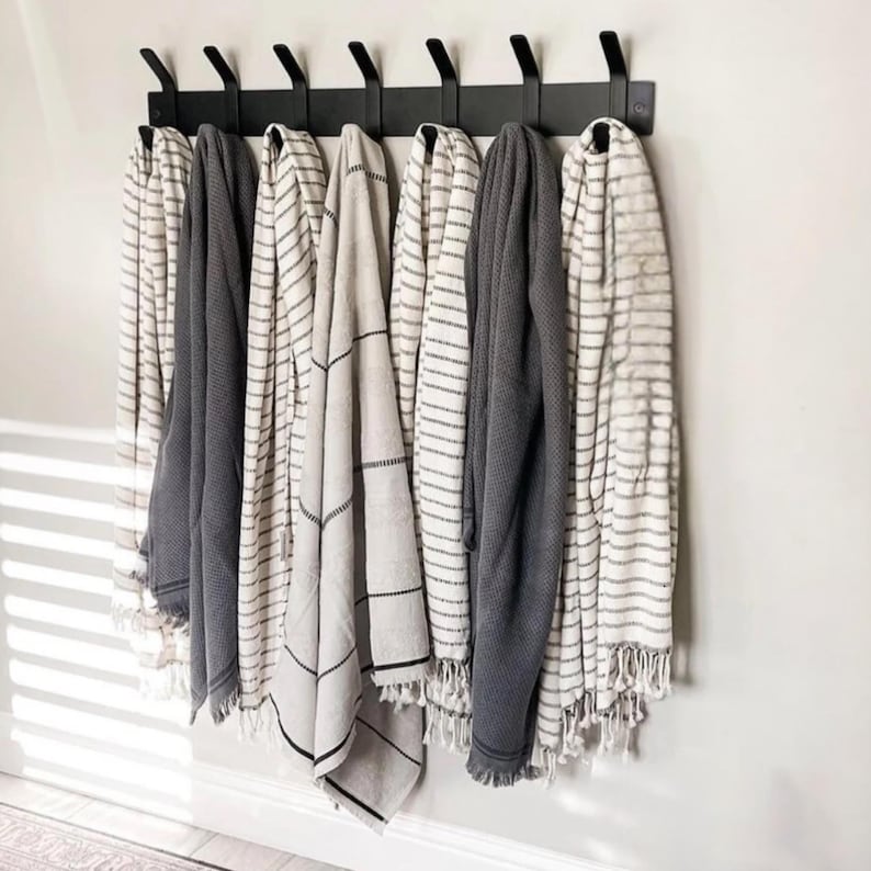 May include: A black metal rack with seven hooks holds a variety of striped and patterned towels. The towels are in shades of white, gray, and black.