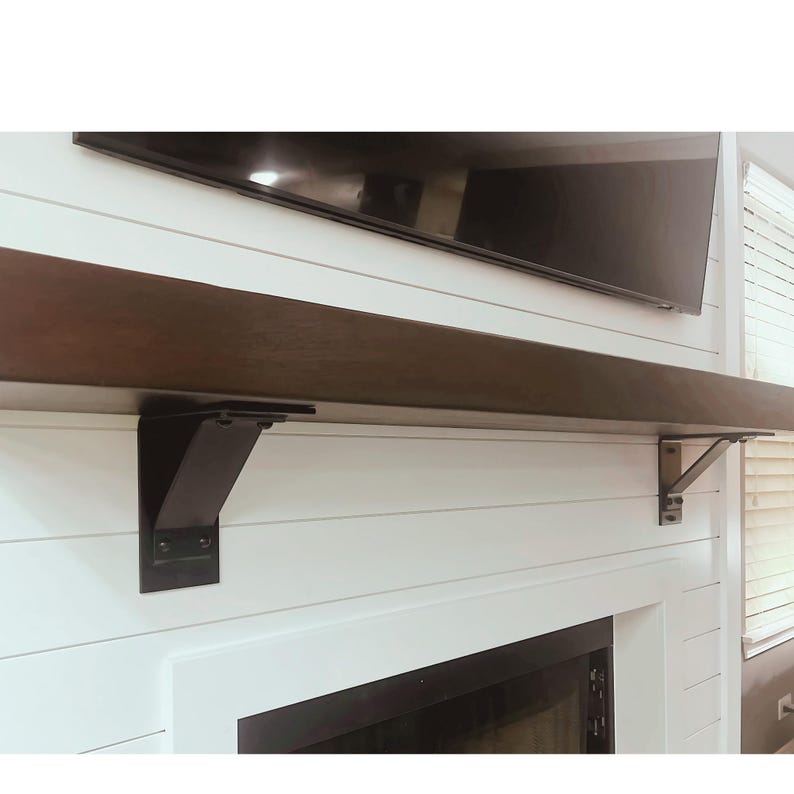 May include: A dark brown wooden shelf with black metal brackets mounted above a fireplace. The shelf is supported by two brackets, each with a decorative design.
