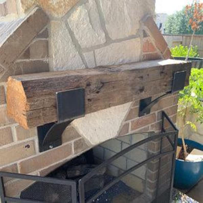 May include: A rustic wooden mantel shelf, approximately 60 inches long, mounted on a brick and stone fireplace. The shelf is supported by black metal brackets. A black metal fireplace screen is in front of the fireplace.