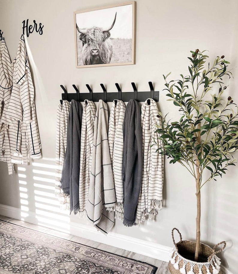 May include: A black metal coat rack with seven hooks mounted on a white wall.  The rack is holding six towels, two white with black stripes, two gray with white stripes, and two gray.  The word "Hers" is written in black on the wall above the rack.  A framed black and white photograph of a cow is hanging on the wall to the right of the rack.  A potted olive tree is on the right side of the image.