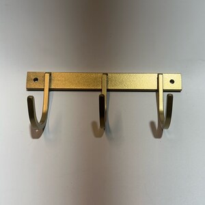 May include: Gold metal wall mounted hook rack with three hooks. The hooks are curved and point downwards.