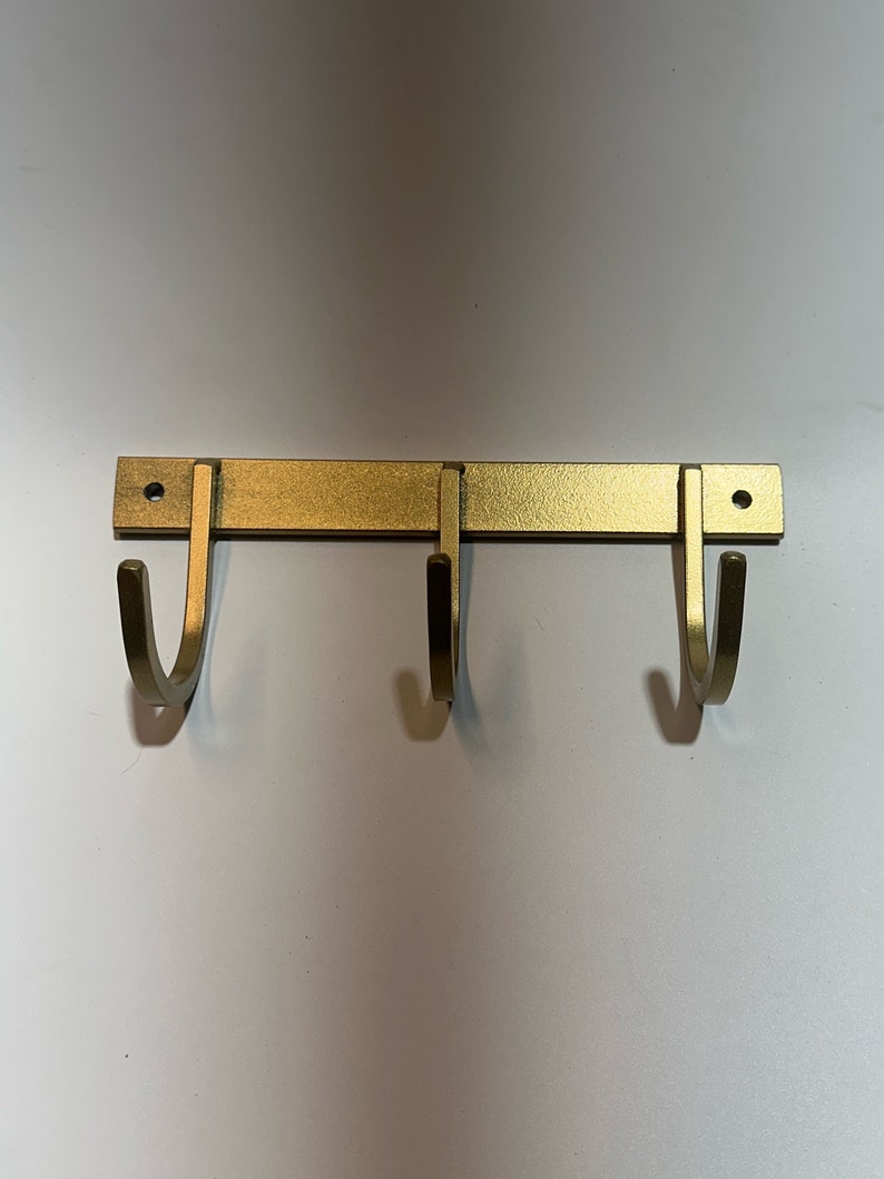 May include: Gold metal wall mounted hook rack with three hooks. The hooks are curved and point downwards.