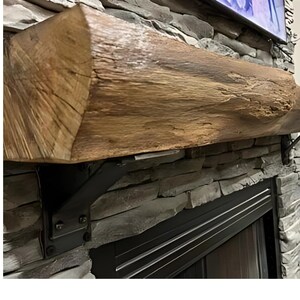May include: A rustic wooden mantel shelf with a dark brown finish, mounted above a black fireplace with a metal grate. The shelf is supported by two black metal brackets.