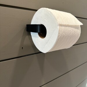 May include: A white paper towel roll is mounted on a black wall-mounted paper towel holder. The holder is attached to a light brown wall.