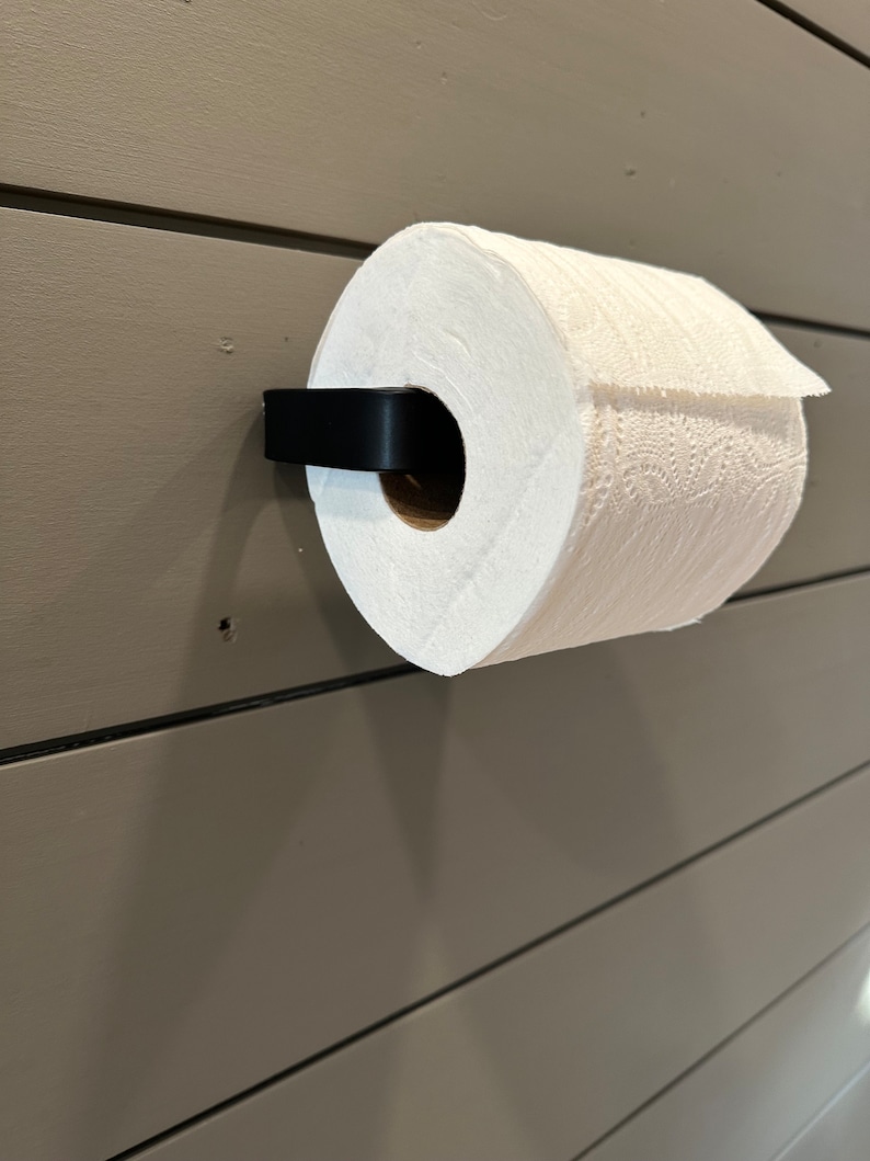 May include: A white paper towel roll is mounted on a black wall-mounted paper towel holder. The holder is attached to a light brown wall.