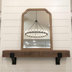 May include: A large, arched mirror with a wooden frame hangs above a rustic wooden mantel. The mirror reflects a chandelier with multiple lights. The mantel is supported by black metal brackets, set against a white shiplap wall.