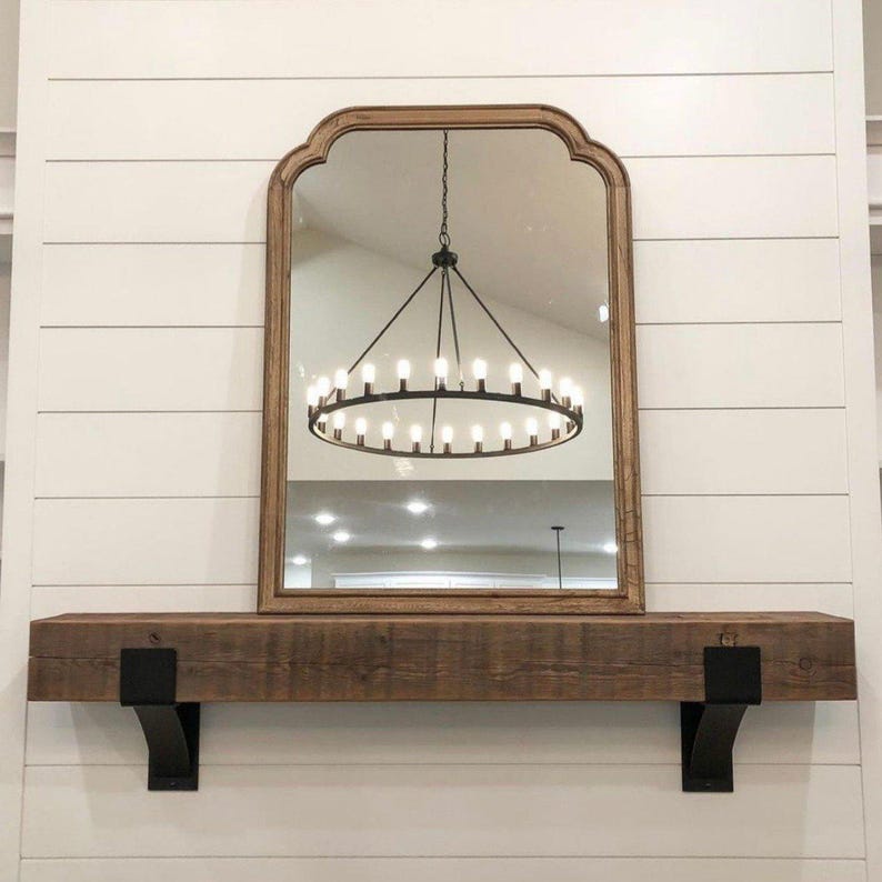 May include: A large, arched mirror with a wooden frame hangs above a rustic wooden mantel. The mirror reflects a chandelier with multiple lights. The mantel is supported by black metal brackets, set against a white shiplap wall.