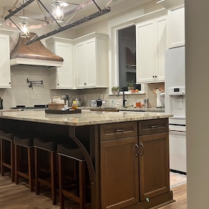 May include: A kitchen island with a granite countertop and brown cabinets. The island has a built-in bar with four stools. The kitchen has white cabinets and a copper range hood.