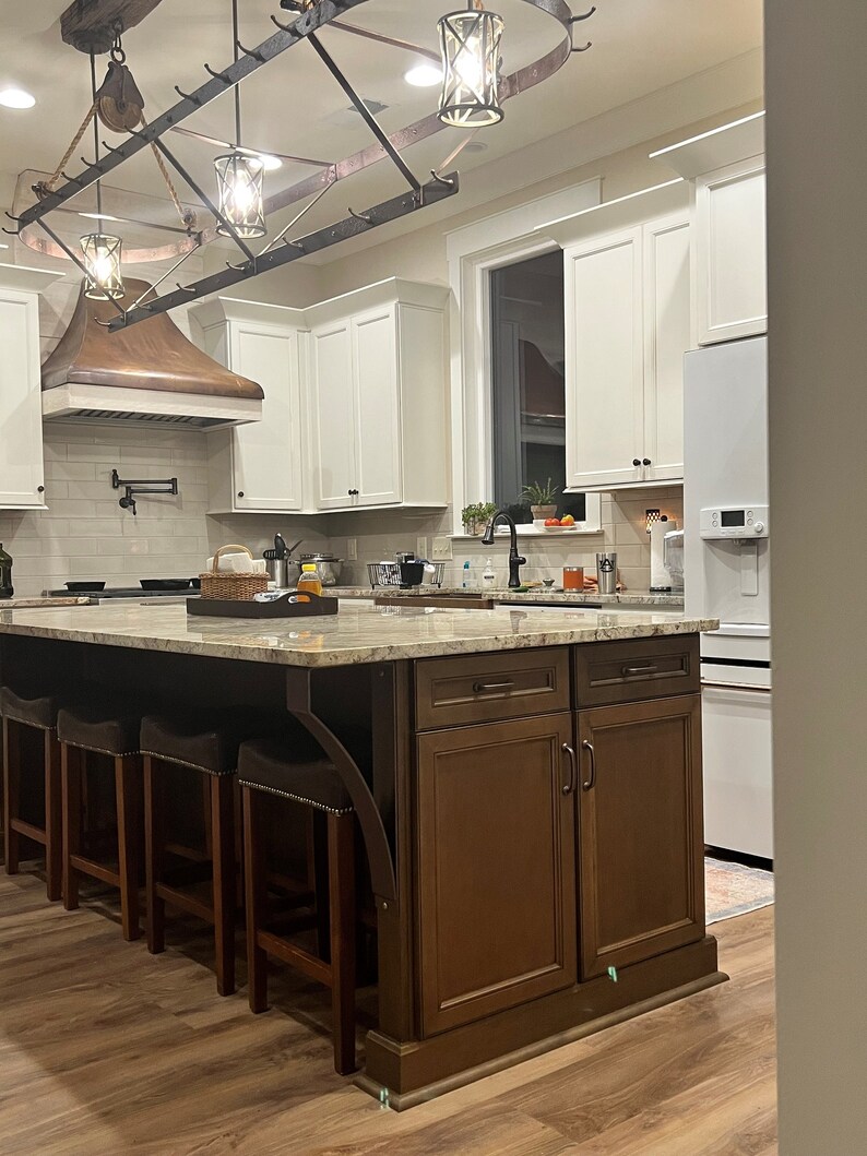 May include: A kitchen island with a granite countertop and brown cabinets. The island has a built-in bar with four stools. The kitchen has white cabinets and a copper range hood.