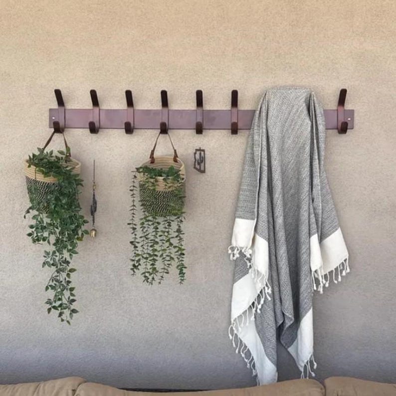 K&ouml;nnte beinhalten: Eine dunkelbraune Wandgarderobe aus Holz mit acht Haken. Zwei geflochtene K&ouml;rbe mit h&auml;ngenden Gr&uuml;npflanzen h&auml;ngen an den Haken. Ein grau-wei&szlig; gestreiftes Textil mit Fransen h&auml;ngt an der rechten Seite der Garderobenleiste.
