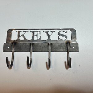 May include: A metal key holder with the word "KEYS" cut out at the top. The key holder has four curved hooks below the word. The metal is a dark gray color and is mounted on a white wall.