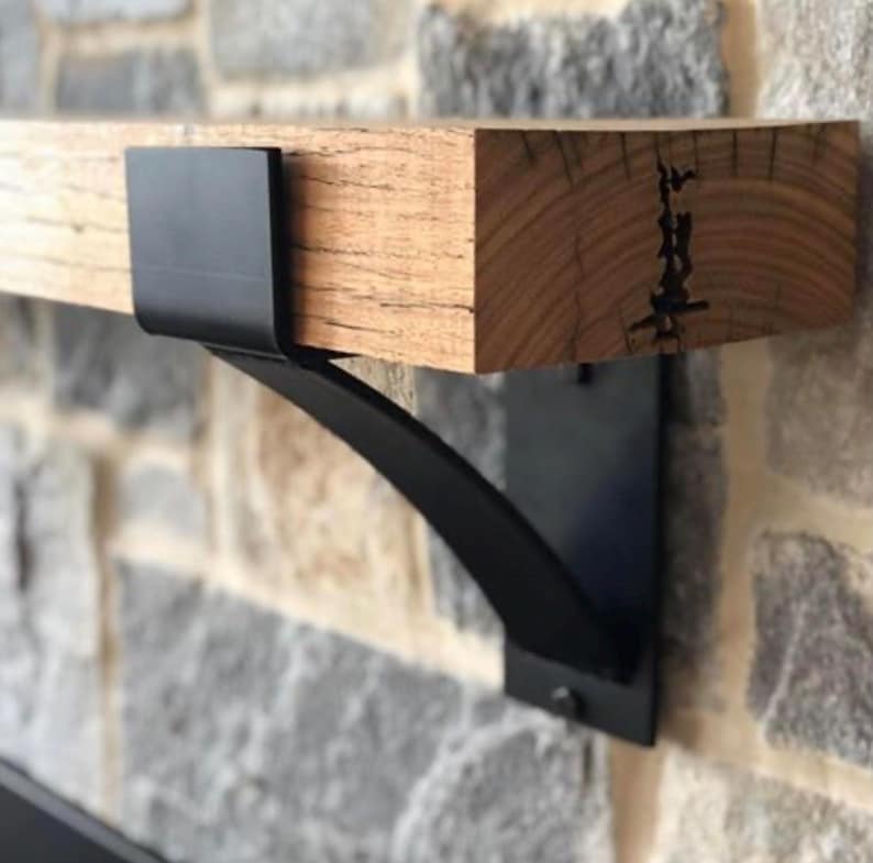 May include: A rustic wooden shelf supported by a black metal bracket. The shelf is made of light brown wood with visible grain and a dark crack. The bracket is a curved design, mounted on a stone wall.