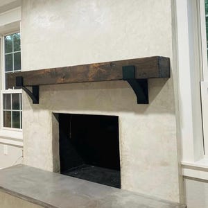 May include: A rustic fireplace with a dark stained wooden mantel supported by black metal brackets. The fireplace has a light-colored textured surround and a dark interior. A concrete hearth extends from the base.