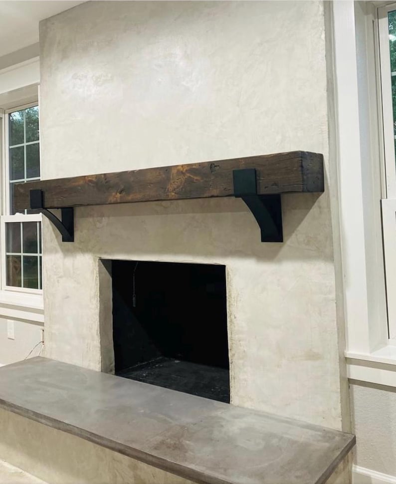 May include: A rustic fireplace with a dark stained wooden mantel supported by black metal brackets. The fireplace has a light-colored textured surround and a dark interior. A concrete hearth extends from the base.