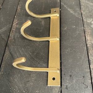 May include: Gold metal wall mounted hook rack with three hooks. The hooks are curved and are attached to a vertical metal bar.