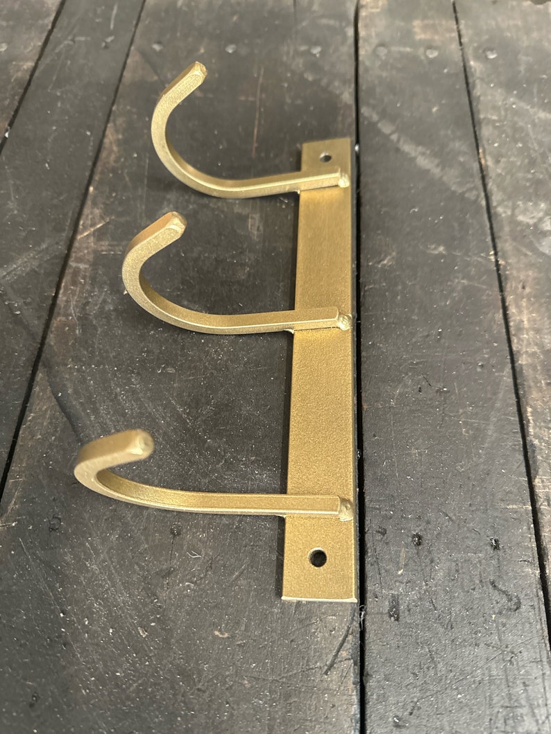 May include: Gold metal wall mounted hook rack with three hooks. The hooks are curved and are attached to a vertical metal bar.