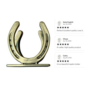 May include: A gold-toned metal napkin holder shaped like a horseshoe, featuring a flat rectangular base and the word CLASSIC embossed on the side. The design includes two parallel horseshoe-shaped frames connected to a stable bottom support piece.