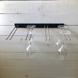 May include: A black metal wine glass rack mounted on a white wall. The rack has four metal rods that hold the glasses upside down. There are two wine glasses hanging on the rack.