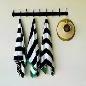 May include: Three black and white striped beach towels with green trim hanging on a black metal rack. A straw hat is hanging on a hook to the right of the towels.