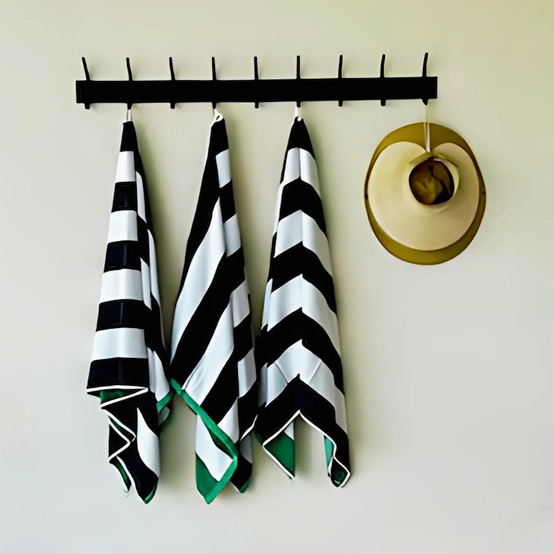 May include: Three black and white striped beach towels with green trim hanging on a black metal rack. A straw hat is hanging on a hook to the right of the towels.