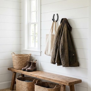 May include: A rustic wooden bench with three woven seagrass baskets underneath, a pair of brown leather boots, a beige canvas tote bag, and a dark olive green waxed jacket hanging on a black horseshoe-shaped wall hook against white shiplap.