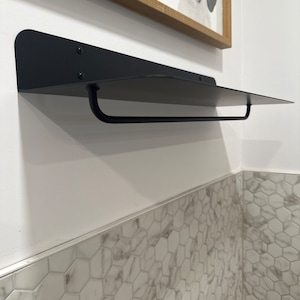 May include: Black metal shelf with a rounded edge and a black metal bar attached to the bottom. The shelf is mounted on a white wall above a white and gray hexagonal tile wall.