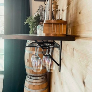 May include: A black metal wine glass rack mounted on a wooden wall with two stacked wooden barrels underneath. The rack holds four wine glasses.