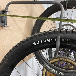 May include: A close-up of a metal bike rack with a tape measure showing 11 inches. The rack is mounted to a wooden wall and holds a black mountain bike tire with the words "BUTCHER GRID TR" printed on the side.