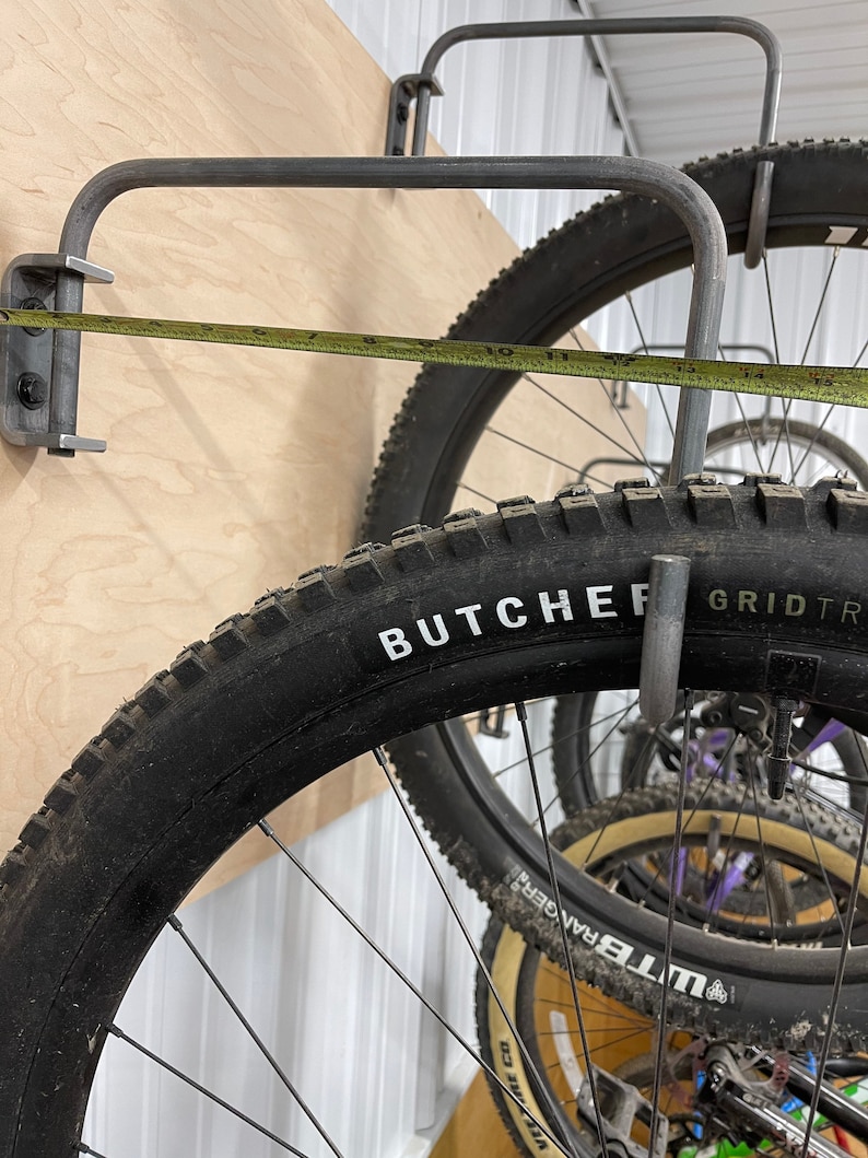 May include: A close-up of a metal bike rack with a tape measure showing 11 inches. The rack is mounted to a wooden wall and holds a black mountain bike tire with the words "BUTCHER GRID TR" printed on the side.