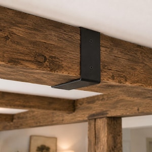 May include: A matte black metal L-shaped bracket secured to a textured, dark brown wooden ceiling beam with two visible screws. The bracket wraps around the corner of the beam, providing a structural connection against the white ceiling surface.