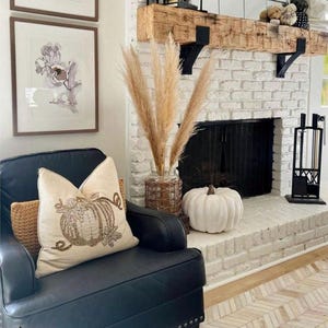 May include: A cozy living room scene with a black leather armchair, a decorative pumpkin pillow, and a white pumpkin on the hearth. A brick fireplace with a wooden mantel and framed floral art complete the autumn-themed decor.