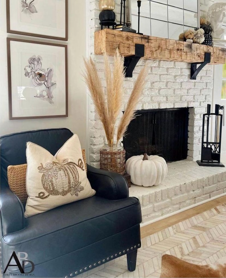 May include: A cozy living room scene with a black leather armchair, a decorative pumpkin pillow, and a white pumpkin on the hearth. A brick fireplace with a wooden mantel and framed floral art complete the autumn-themed decor.