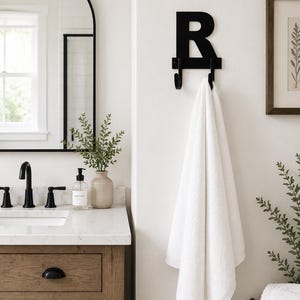 May include: A black metal wall-mounted hook shaped like the capital letter R, featuring two curved prongs at the bottom. The hook is mounted on a white wall and holds a single white towel, providing a functional and decorative storage solution.