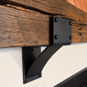 May include: A matte black metal shelf bracket with a curved support arm and a flat mounting plate. The bracket is secured to a wall and supports a thick, weathered brown wooden beam, featuring four visible circular rivets on the top plate.