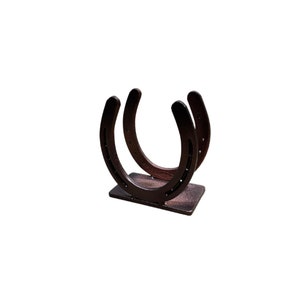 May include: A dark brown metal horseshoe holder featuring two U-shaped horseshoes welded vertically onto a rectangular base. The horseshoes are positioned parallel to each other, creating a cradle-like structure with visible nail holes along the rims.