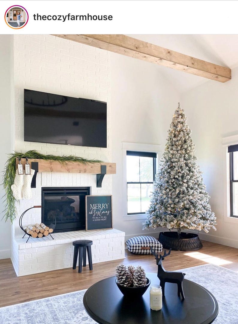 May include: A cozy living room decorated for Christmas. A large, flocked Christmas tree stands next to a fireplace with stockings. A black coffee table holds pine cones and a candle. A sign reads "Merry Christmas."
