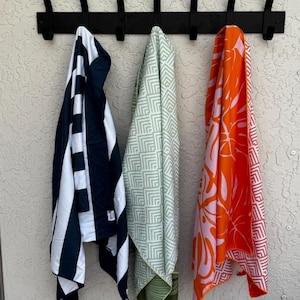 K&ouml;nnte beinhalten: Eine schwarze Wandgarderobe mit sieben Haken h&auml;lt drei h&auml;ngende Handt&uuml;cher. Das linke Handtuch hat marineblaue und wei&szlig;e vertikale Streifen. Das mittlere Handtuch zeigt ein hellgr&uuml;nes und wei&szlig;es geometrisches Muster. Das rechte Handtuch hat einen orangefarbenen und hellrosa tropischen Blattdruck.